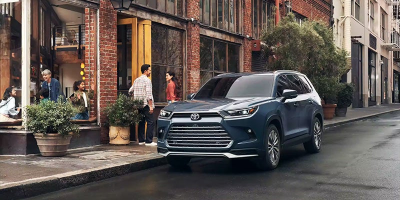 An all-new Storm Cloud Blue 2024 Toyota Grand Highlander Hybrid parked on a city street next to a couple walking into a brick building. - Phil Meador Toyota Dealership Pocatello, ID